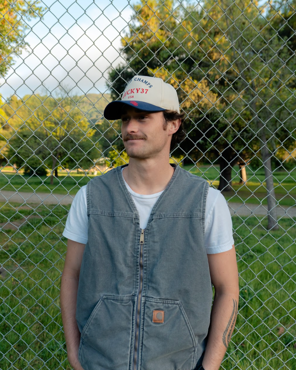 Person wearing a cap and vest standing behind a chain-link fence with trees in the background.
