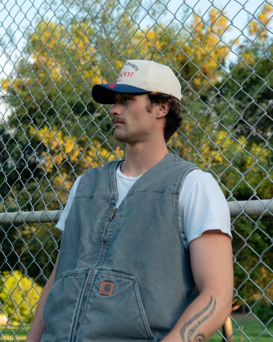Man wearing a cap and vest standing behind a chain-link fence.