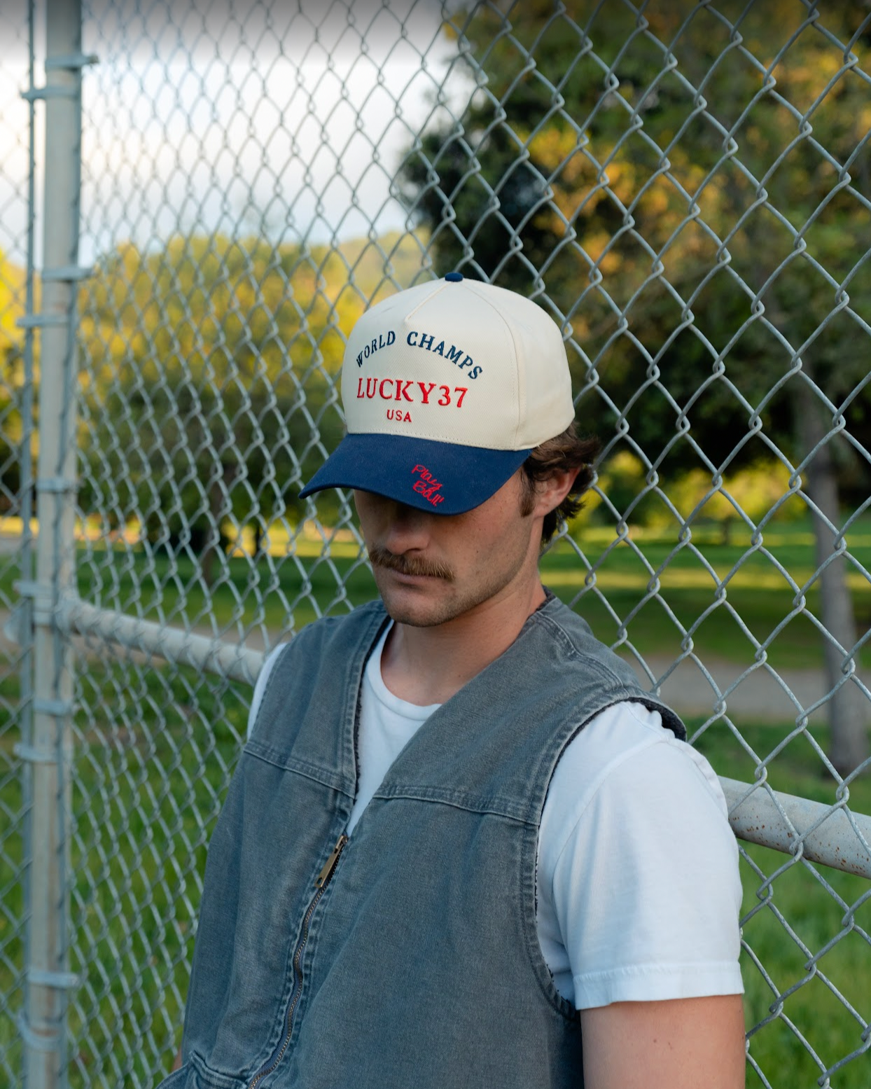 A person wearing a navy trucker cap with the text 'WORLD CHAMPS LUCKY37 USA' on it, standing behind a chain-link fence.