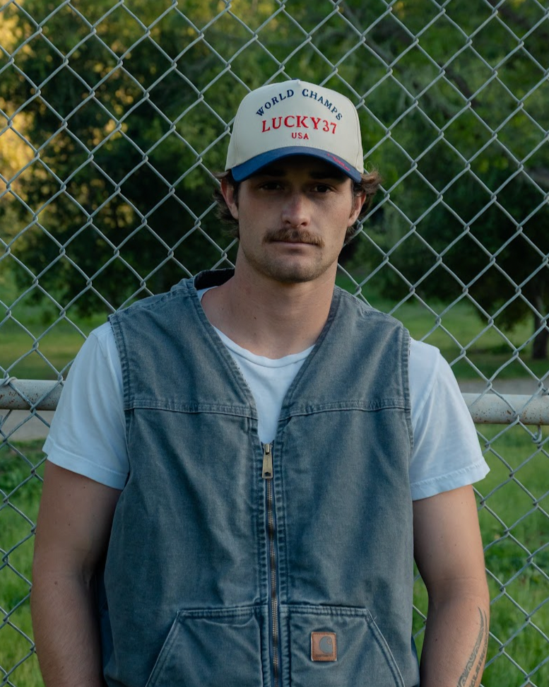 Man wearing a cap and vest standing in front of a chain-link fence.